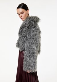Grå faux fur cropped jakke med lange ærmer og hætte. Båret sammen med en bordeauxfarvet plissee nederdel, der fremhæver en tekstureret lagdeling.