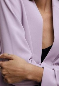 Blazer lavande clair à la texture lisse et aux manches larges, porté sur un haut noir, avec un devant ouvert et un design minimaliste.