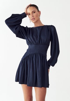Femme portant une robe bleu marine à manches longues bouffantes et taille haute, posant avec une main sur la tête devant un fond blanc.