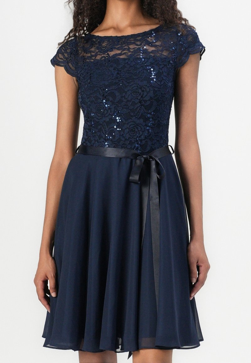 Robe bleu marine avec un corsage en dentelle orné de sequins, des manches courtes, une jupe évasée en chiffon et une ceinture en ruban de satin à la taille.