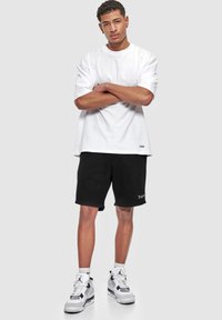 Weißes Baumwoll-T-Shirt, lockere Passform, kombiniert mit schwarzen Baumwollshorts mit gesticktem Logo. Das Modell trägt graue und schwarze Sneakers mit weißen Socken.