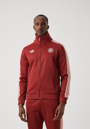 Red athletic zip-up jacket featuring white stripes on the sleeves, a high collar, and a circular logo on the chest. Smooth fabric texture.
