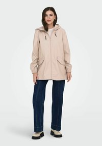 Imperméable beige claro con capucha, cierre frontal, puños elásticos y dos bolsillos laterales, combinado con unos vaqueros de denim oscuros y zapatos de slip-on.