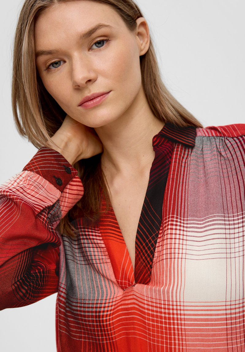 Camicia in flanella a quadretti rossi e neri con scollatura a V, maniche lunghe e polsini con bottoni. Tessuto leggero con un motivo a griglia e una texture morbida.