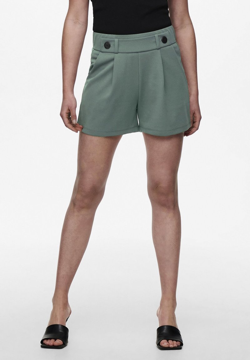 Pantaloni corti plissettati verdi con un alto elastico in vita, dotati di due bottoni neri. Tessuto liscio, lunghezza al ginocchio, abbinati a sandali neri a scivolo.