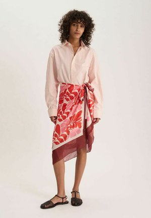 Femme aux cheveux bouclés portant une chemise rayée rose pâle rentrée dans une jupe portefeuille rouge et rose à fleurs, avec des sandales noires ouvertes au bout des orteils.