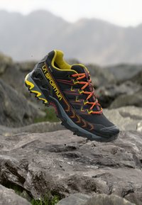 Črne tekaške čevlje za trail z rumenimi poudarki, rdečimi vezalkami in teksturiranim podplatom. Imajo podporno zasnovo in ojačano kapico prstov.