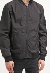 Black bomber jacket with a front zipper, ribbed cuffs and hem, side pockets, and a zippered sleeve pocket. Smooth, lightweight material.