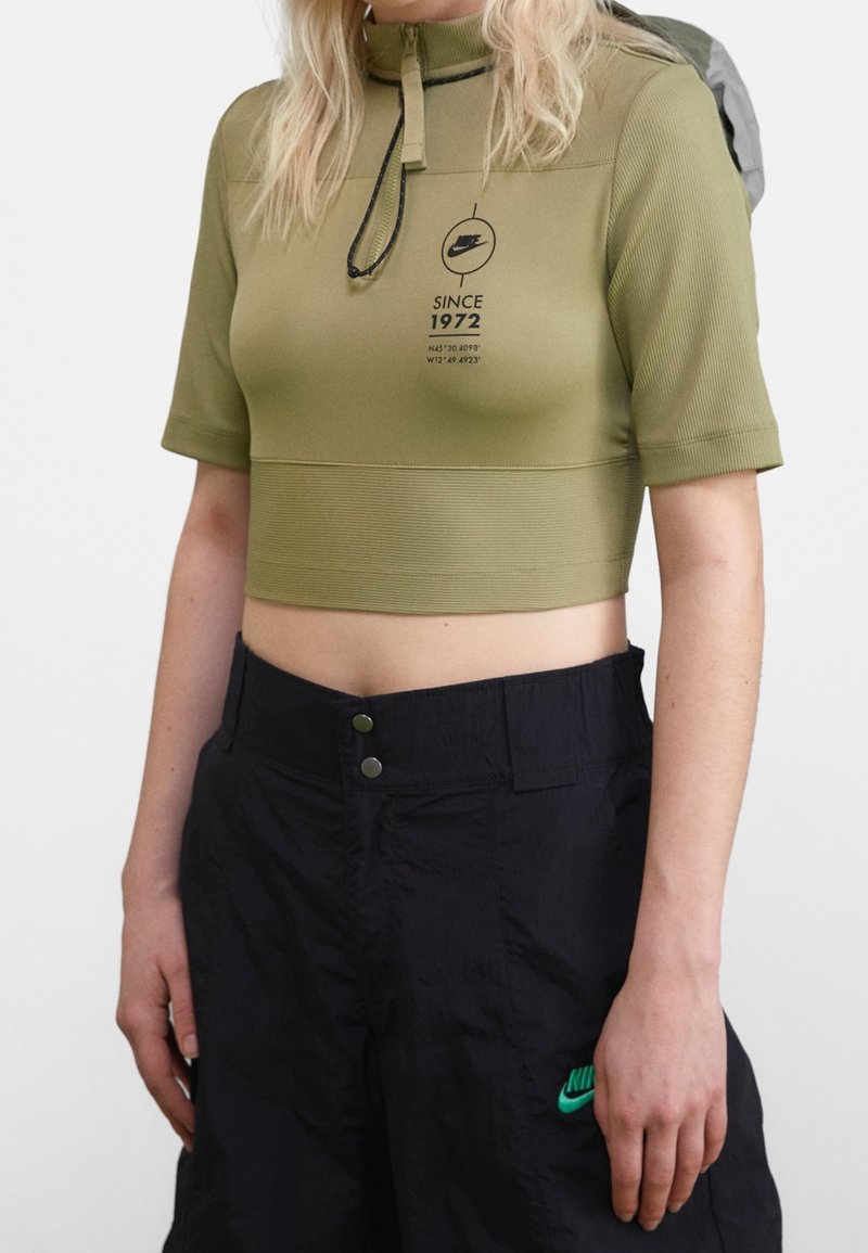 Olivově zelený zkrácený top s krátkými rukávy, vysokým límcem, zipem a logem. V kombinaci s černými kalhotami s zeleným logem a cvokovými knoflíky.
