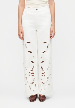 Γυναίκα με λευκό παντελόνι φαρδιάς γραμμής με συμμετρικά σχέδια λουλουδιών με κοψίματα και καφέ παπούτσια με τετράγωνη μύτη, που στέκεται μπροστά σε απλό φόντο.