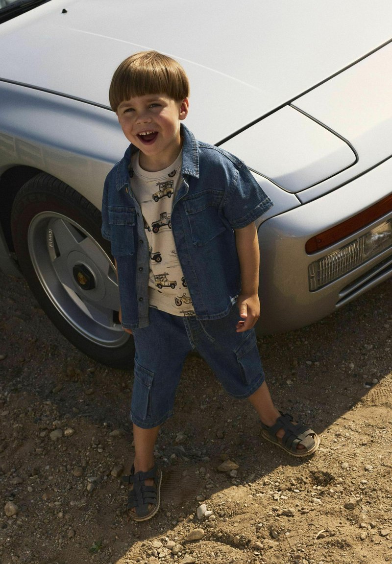 Giacca di jeans, t-shirt grafica con design di veicolo e pantaloncini di jeans. In piedi vicino a un'auto argentata su una superficie sabbiosa. Sandali marroni.