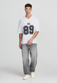 Biała koszulka o oversize'owym kroju z dekoltem w serek, z czarno-białą grafiką z numerem 89. W zestawieniu z jasnoszarymi dżinsami i białymi sneakerami.