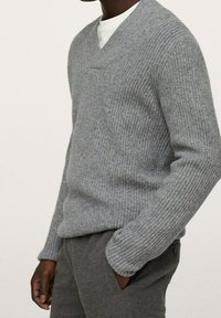 Grauer gestrickter Pullover mit V-Ausschnitt, gerippter Textur und einer Brusttasche. Kombiniert mit grauen Hosen, die einen bequemen, lässigen Stil zeigen.