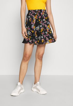 Kobieta ubrana w żółty top bez rękawów, czarną kwiecistą spódnicę i białe masywne sneakersy, stojąca na tle jasnego, jednolitego tła.