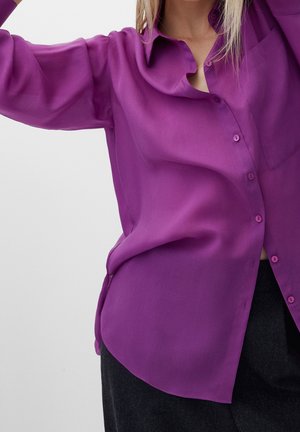 Femme portant une chemise ample, violet vif, boutonnée avec manches longues et un pantalon foncé, sur un fond clair uni.