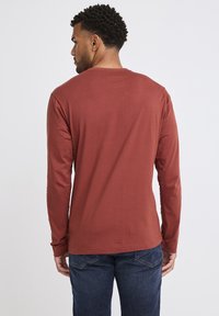 Lange mouw, aardse rode t-shirt met een ronde hals en een gladde textuur, gecombineerd met donkerblauwe jeans. Eenvoudig achteraanzicht, zonder patronen of accenten.