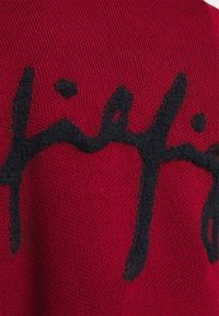 Czerwony sweter z dzianiny z wyraźnym czarnym, puszystym wzorem logo w formie napisu. Tekstura wydaje się miękka, a krój jest dopasowany.