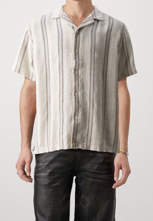 Mand iført cream kortærmet skjorte med lodrette blå striber og sorte teksturerede bukser, med perlearmbånd på højre håndled.