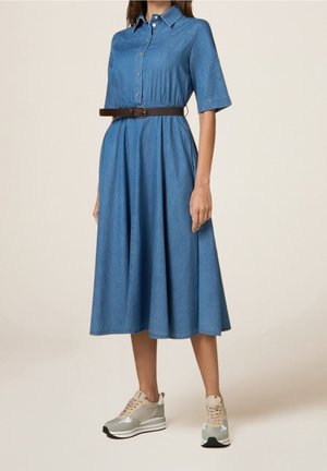 Kobieta w długiej do połowy uda sukience z denimu z krótkimi rękawami, zapinanej na guziki z przodu, z brązowym paskiem i beżowymi sneakersami na platformie, stojąca na jednolitym tle.