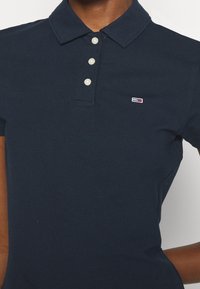 Polo bleu marine avec trois boutons blancs et un petit logo rectangulaire rouge, blanc et bleu sur la poitrine gauche.
