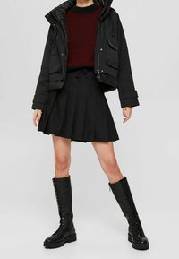 Doudoune noire avec doublure matelassée, pull en tricot rouge, jupe plissée noire et bottes noires à lacets montantes.