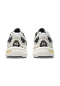 Pari kengät urheilullisia harmaa-ja-valkoisia värejä mustilla verkkopaneeleilla ja "GEL"-merkillä kantapäässä, näkyvät takaa valkoista taustaa vasten.