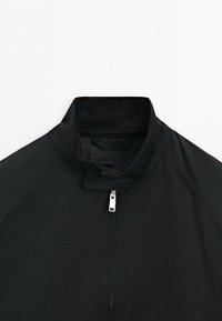 Black jacket with a raised collar and silver zipper, showing detailed stitching around the shoulders and collar area.