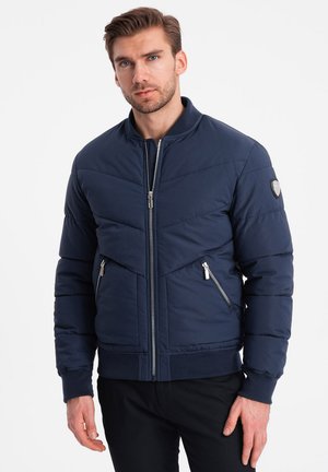 Marineblaue Daunenjacke mit Reißverschluss vorne, gerippten Bündchen und zwei seitlichen Reißverschlusstaschen. Mit einem Chevron-Nähmuster und einem Logo-Patch am Ärmel.