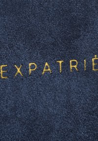 Marinblå mocka med präglad guldtext som stavas "EXPATRIÉ". Har en slät, mjuk yta och en minimalistisk design utan ytterligare accenter.