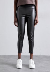 Person som bär svarta byxor i konstläder och vita sneakers, ståendes mot en grå bakgrund av vägg och golv.