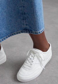 White sneaker with textured leather upper, round toe, and flat rubber sole. It features flat laces and subtle stitching details.