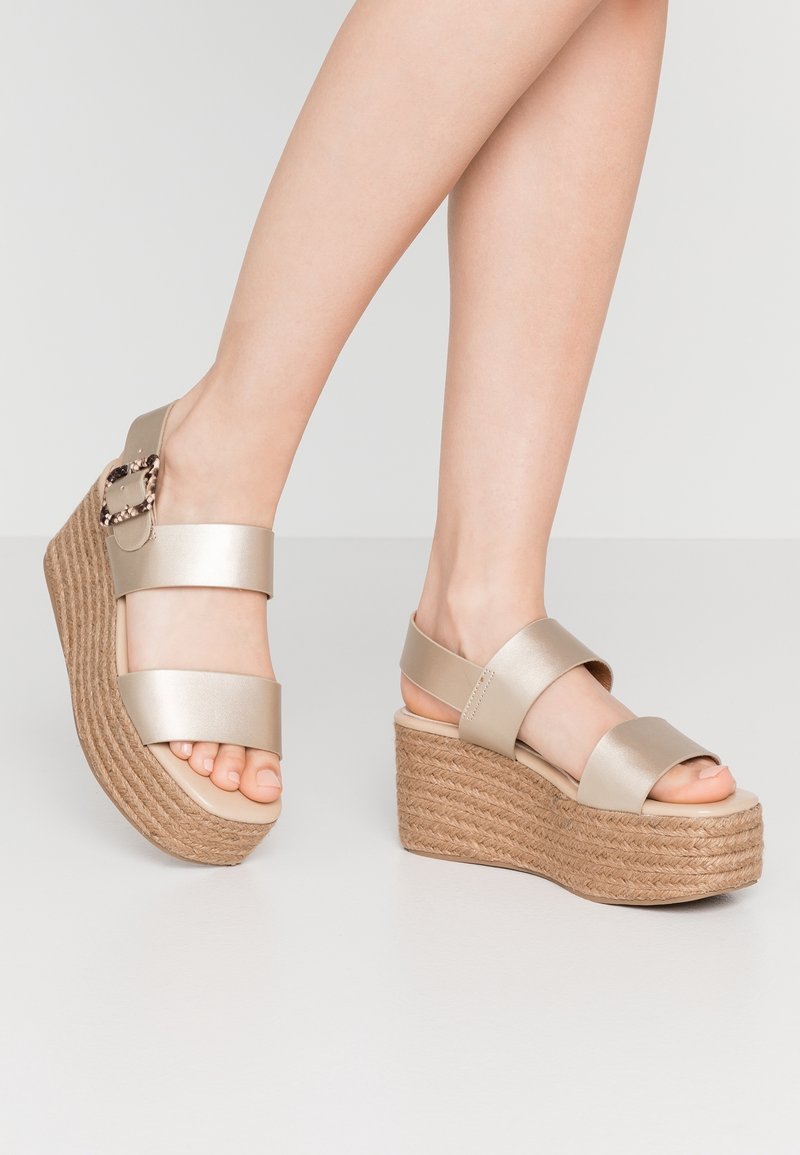 Sandales compensées beiges avec deux larges brides, un talon en corde texturée et un détail de boucle décorative. Matériau lisse avec un design à bout ouvert.