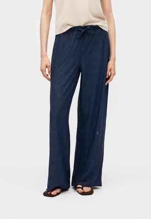 Persona che indossa una canotta beige, pantaloni larghi blu navy con motivi floreali traforati e infradito nere, in piedi su pavimento bianco.