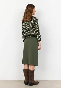 Haut à manches longues avec un motif géométrique noir et crème, associé à une jupe vert olive longueur genou et des bottes en cuir marron au niveau de la cheville.