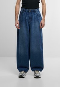 Pantaloni in denim blu a gamba larga con vita alta, dotati di chiusura a bottone, tasche laterali e una texture sbiadita. Abbinati a sneakers grigie.