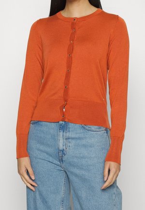 Femme portant un cardigan boutonné orange brûlé avec des poignets et un ourlet côtelés, associé à un jean bleu clair à taille haute.