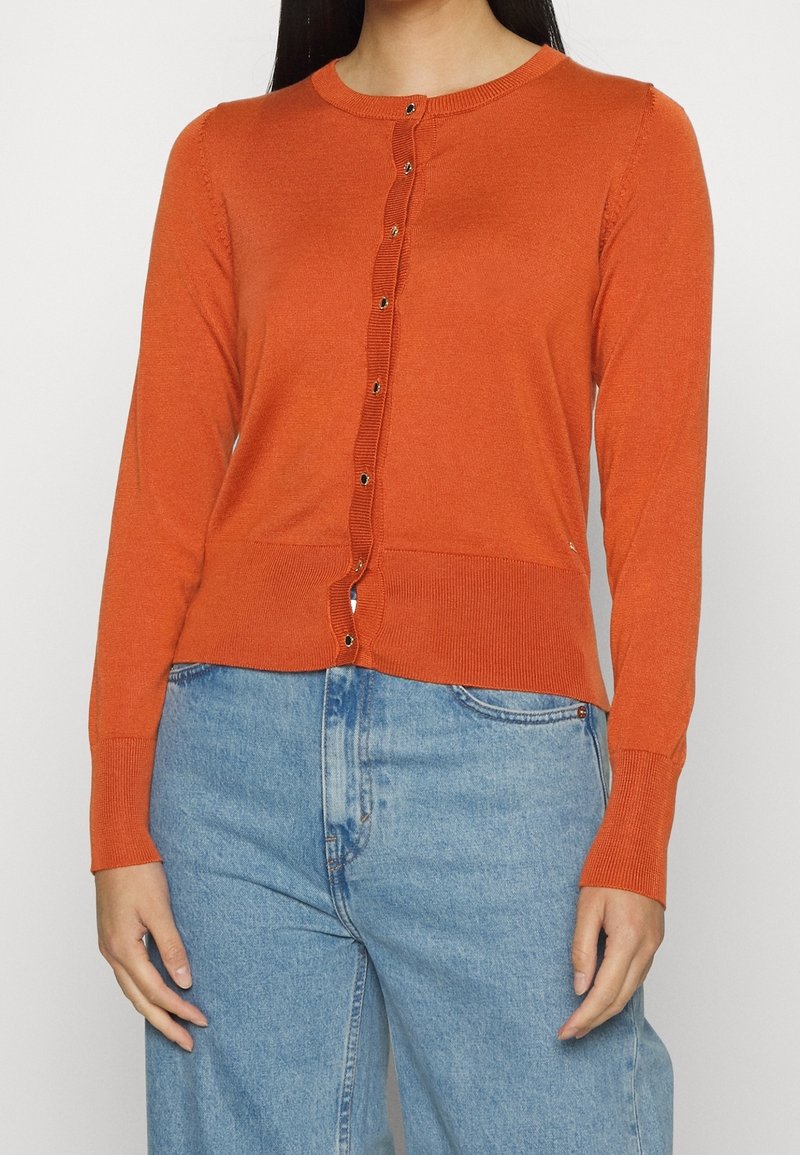 Femme portant un cardigan boutonné orange brûlé avec des poignets et un ourlet côtelés, associé à un jean bleu clair à taille haute.