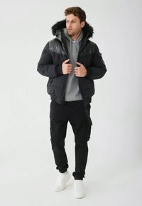 Mężczyzna w czarnej kurtce puchowej z futrzanym kapturem, pod którą ma szary hoodie, czarne spodnie cargo i białe sneakersy, stojący na jednolitym tle.