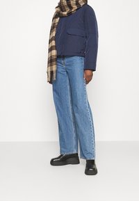 Person som bär en marinblå jacka, blå jeans, svarta plattformsstövlar och en beige- och brunt rutig sjal mot en enfärgad bakgrund.