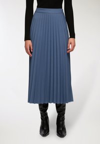 Gonna lunga plissettata in un morbido tessuto blu. Presenta un alto cinturino in vita e arriva fino a metà polpaccio, abbinata a stivali neri attillati fino al ginocchio.