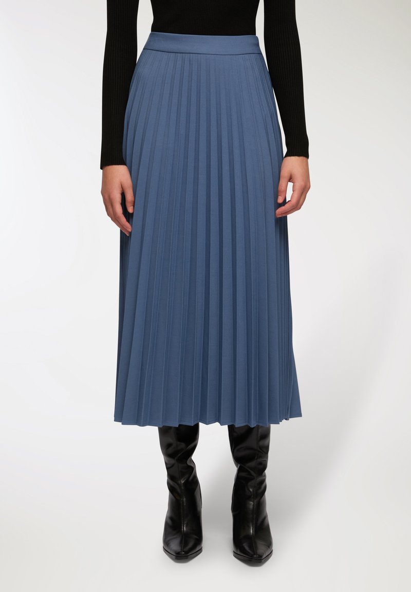 Gonna lunga plissettata in un morbido tessuto blu. Presenta un alto cinturino in vita e arriva fino a metà polpaccio, abbinata a stivali neri attillati fino al ginocchio.