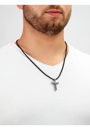 Schwarze geflochtene Halskette mit einem metallischen silbernen Kreuzanhänger, getragen über einem schlichten weißen Hemd. Das Kreuz verfügt über eine polierte Oberfläche und geometrische Details.