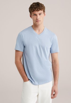 Jonge man met krullend haar, gekleed in een lichtblauw V-hals T-shirt en witte broek, staand met één hand in zijn zak tegen een neutrale achtergrond.