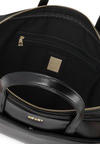 Interno di una borsa a tracolla in pelle nera con texture liscia, dettagli della zip dorati, una tasca con zip e un'etichetta del marchio in design quadrato.