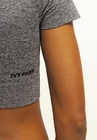 Top corto de manga corta en gris, hecho de tejido de punto, con un logo "IVY PARK" en negro en el lado inferior, textura suave y diseño ajustado.