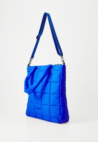 Sac fourre-tout bleu matelassé avec une texture lisse, présentant un design rembourré, une bandoulière et un détail de logo. Fermeture éclair en haut.