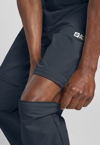 Pantaloni impermeabili grigio scuro con una texture liscia, dotati di una patch con logo e un design aderente, regolati all'altezza del ginocchio.