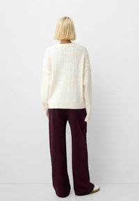 Chunky white cable-knit sweater with a round neck and ribbed hem, paired with loose-fitting, dark burgundy trousers and white shoes.