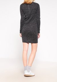 Abito lungo grigio scuro a maniche lunghe con design aderente e zip sul retro. Il modello indossa sneaker bianche e calzini grigi. Tessuto morbido e testurizzato.
