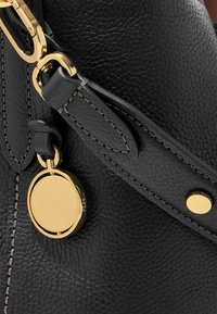 Schwarze Lederhandtasche mit strukturierten Oberflächen, ausgestattet mit goldfarbenen Beschlägen, einem runden Anhänger und kontrastierenden Nähten am Tragegurt.
